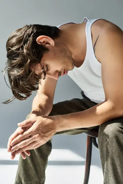 Un joven con un tatuaje se sienta pensativamente en una camiseta sin mangas, reflejándose en un entorno minimalista. — Stock Photo