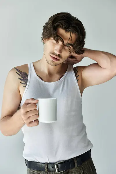 Un joven con estilo en una camiseta sin mangas sostiene una taza blanca mientras se relaja sobre un fondo neutro. - foto de stock