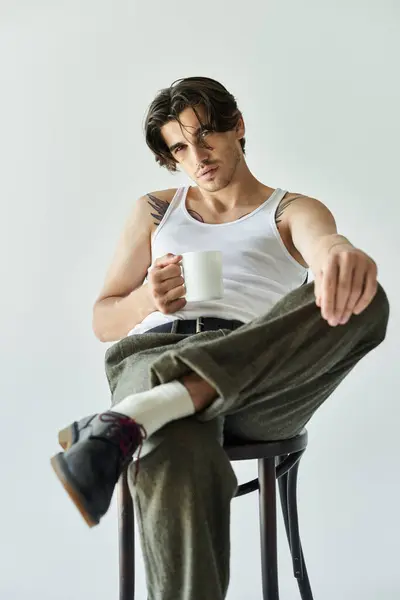 A young man confidently poses on a stool, wearing a tank top, showcasing his tattoo while holding a cup. — Stock Photo