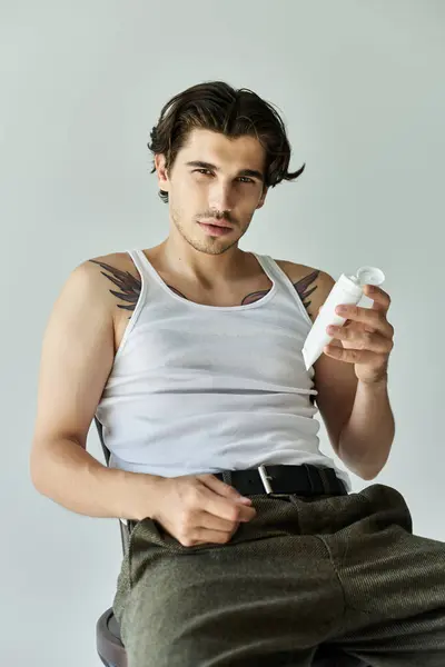 A handsome young man with tattoos relaxes in a tank top, confidently holding a white bottle while posing. — Stock Photo