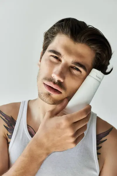 A stylish young man in a tank top leans against a hair product, showcasing his tattoos on a grey backdrop. — Stock Photo