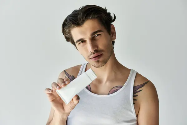A confident young man with striking tattoos displays a skincare product, exuding charm and style in a gray setting. — Stock Photo