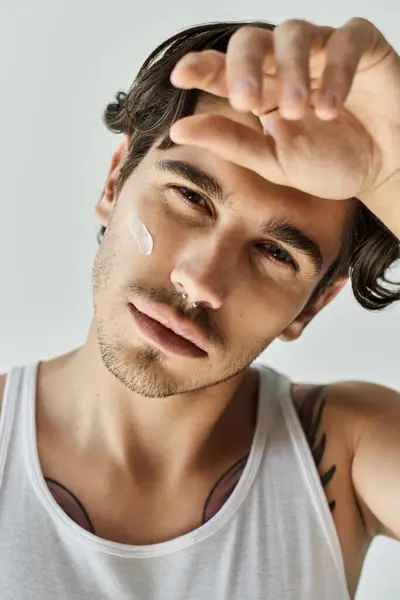 The young man with a trendy tattoo wears a white tank top, gazing intensely while applying cream. — Stock Photo