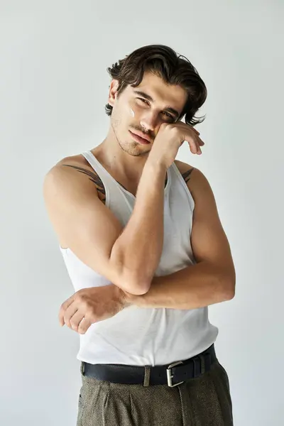 A young man in a tank top shows off his tattoos while striking a contemplative pose against a smooth grey backdrop. — Stock Photo