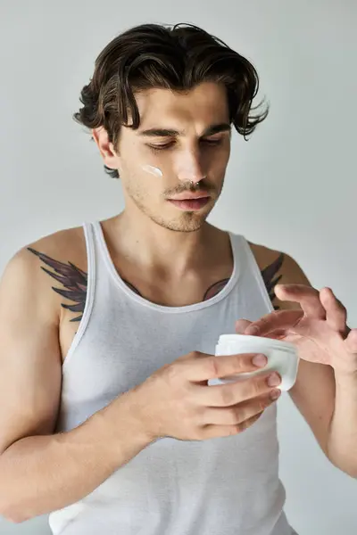 Un jeune homme en débardeur applique de la crème pour la peau, exhibant son tatouage sur un fond gris. — Photo de stock