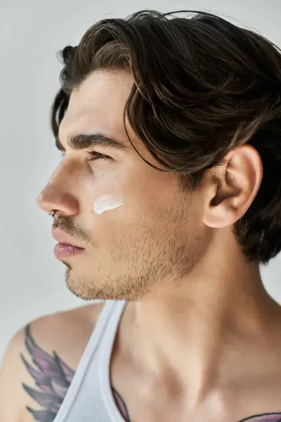 A stylish man in a tank top carefully applies cream to his face, revealing his unique tattoos and focused expression. — Stock Photo