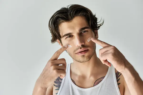 Un bel giovane uomo con un tatuaggio mostra la sua routine di cura della pelle, trasudando fiducia in un ambiente grigio. — Foto stock