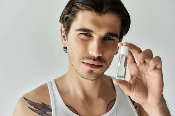 A young handsome man in a tank top confidently presents a small bottle, highlighting his tattoos and style. — Stock Photo
