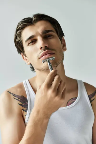 A young man in a tank top contemplates while shaving, showcasing his tattoos against a neutral grey background. — Stock Photo
