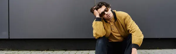 A young man showcases his trendy autumn style while casually sitting against a sleek urban wall outdoors. — Stock Photo