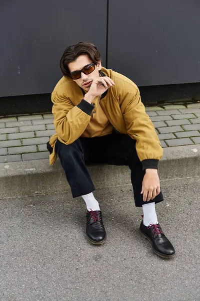 A young, handsome man dons a trendy autumn outfit while sitting on a low wall, showcasing his unique style. — Stock Photo