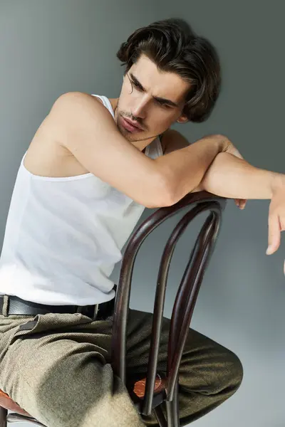 A young, tattooed man leans thoughtfully on a chair, dressed simply in a tank top, exuding relaxed confidence. — Stock Photo