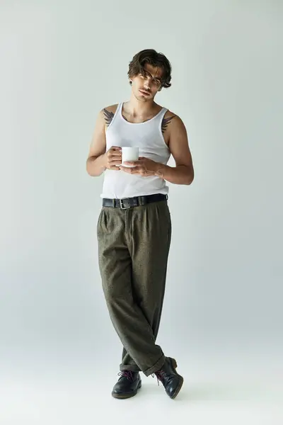 A young man with a tattoo stands confidently, holding a cup while dressed in a tank top and relaxed trousers. — Stock Photo