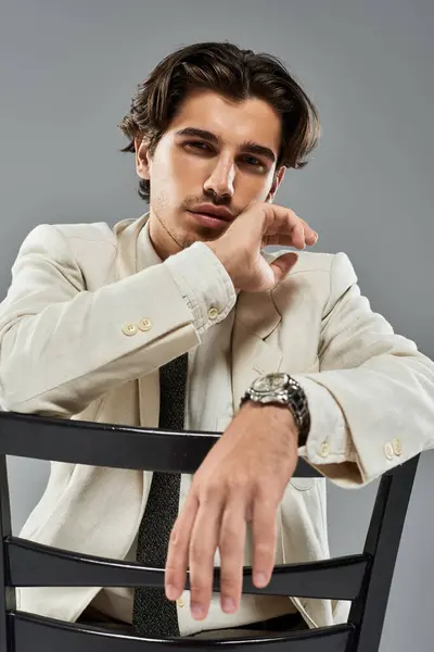 A young, handsome man in formal attire confidently poses with a chair, showcasing his style against a grey background. — Stock Photo