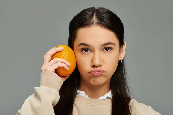 A pretty young woman in stylish attire holds an orange beside her face while expressing emotion. — Foto stock