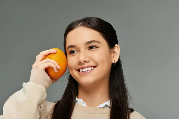 A cheerful young woman in a cozy outfit displays an orange with a radiant smile, embodying joy. — Stock Photo