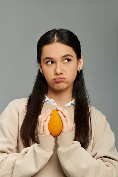 A young woman with long hair holds an orange, displaying a thoughtful and emotional expression. — Stock Photo