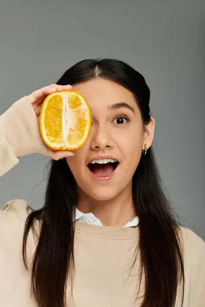 Emotional young woman in stylish attire playfully surprises with a slice of orange, expressing joy. — Stock Photo