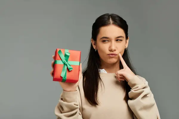 Una joven de moda sostiene un regalo envuelto, meditando alegremente con una expresión lúdica. - foto de stock