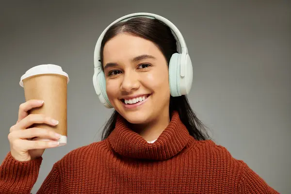 A young woman with a joyful expression holds a coffee cup while wearing headphones. — Stock Photo