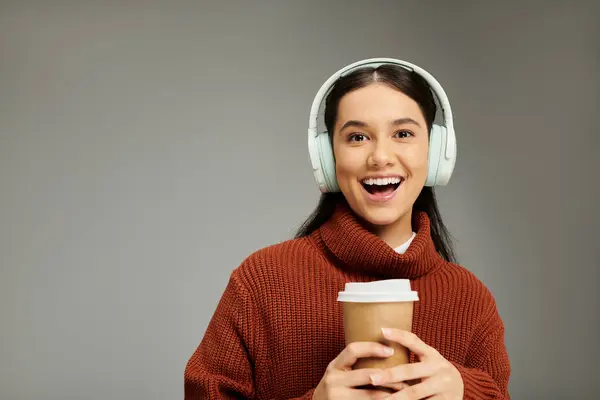 Una giovane donna gioiosa tiene una tazza di caffè e indossa cuffie, irradiando calore e stile. — Foto stock