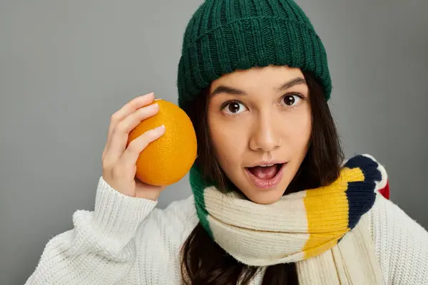 This cheerful young woman expresses surprise while holding an orange, dressed warmly for winter. — Stock Photo