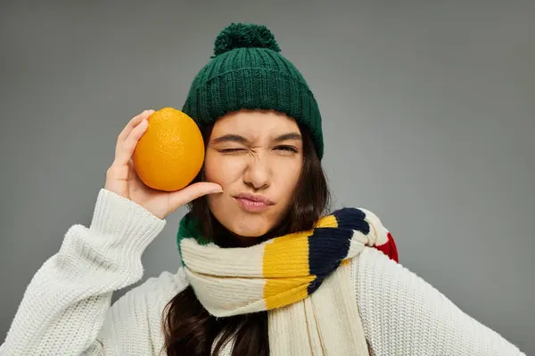 Expressing joy, a stylish young woman winks while holding an orange beside her cheek. — Stock Photo