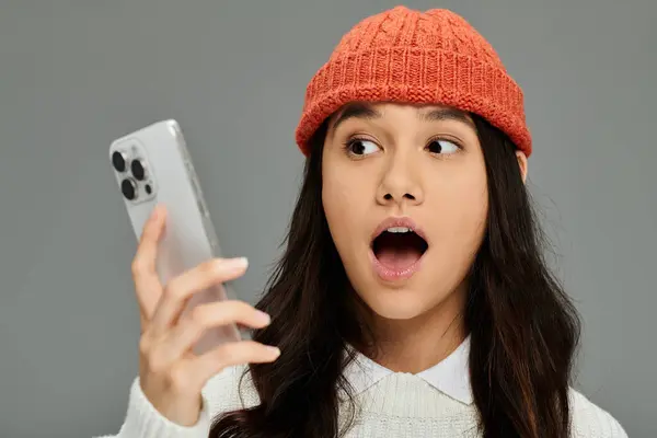 A young woman, wearing a vibrant orange beanie, reacts with surprise as she looks at her phone. — Stock Photo