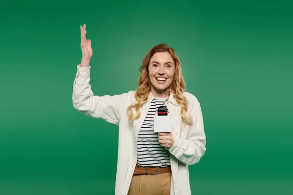 A cheerful woman with curly hair greets with a wave while holding a cup, set against a bright green backdrop. — Stock Photo
