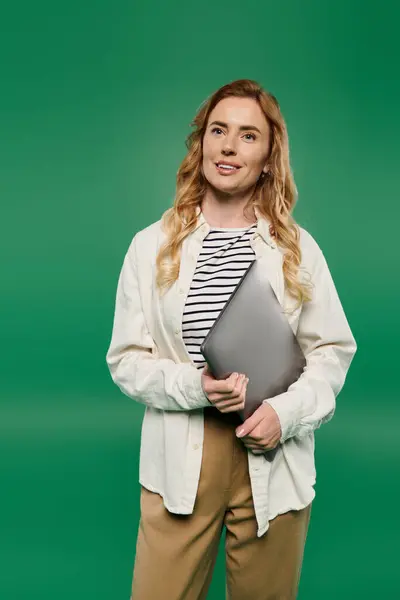 A woman with curly hair in casual attire smiles while holding a laptop, set against a bright green backdrop. — Stock Photo