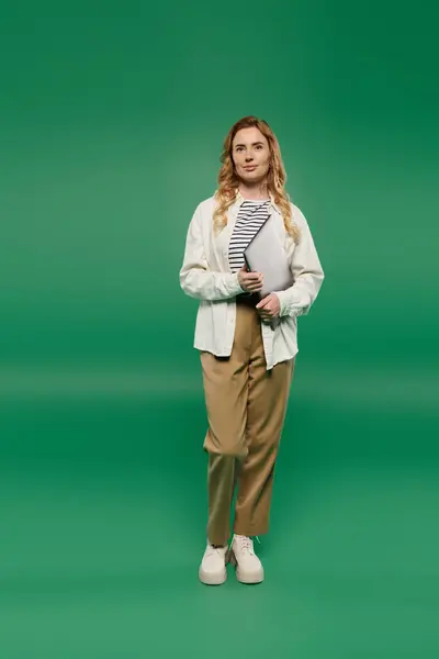 A stylish woman with curly hair poses casually, holding a laptop while surrounded by lush green color. — Stock Photo