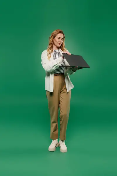 A woman with curly hair wears casual clothing, engaging with a laptop on a striking green backdrop. — Stock Photo