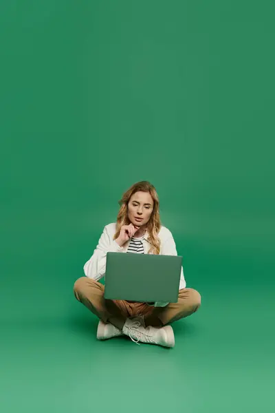 A thoughtful woman with curly hair sits cross-legged, engaged with her laptop in a lively green setting. — Stock Photo