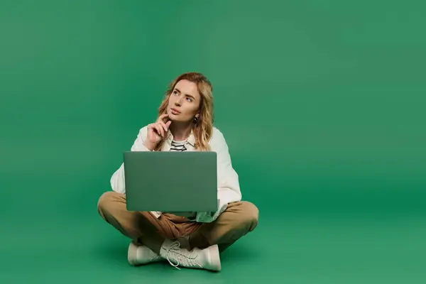 A woman with curly hair sits cross-legged, deep in thought as she uses her laptop on a striking green backdrop. — Stock Photo