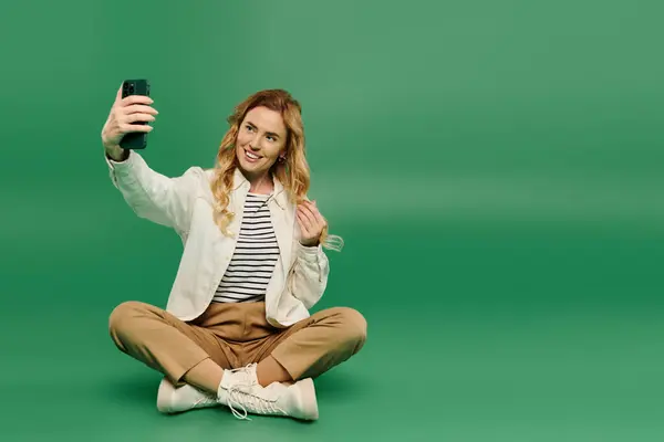 A cheerful woman dressed casually sits cross-legged, enjoying a selfie moment on a vivid green background. — Stock Photo