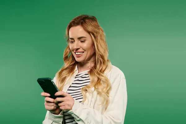 A smiling woman in casual attire interacts with her smartphone, radiating joy on a lively green background. — Stock Photo