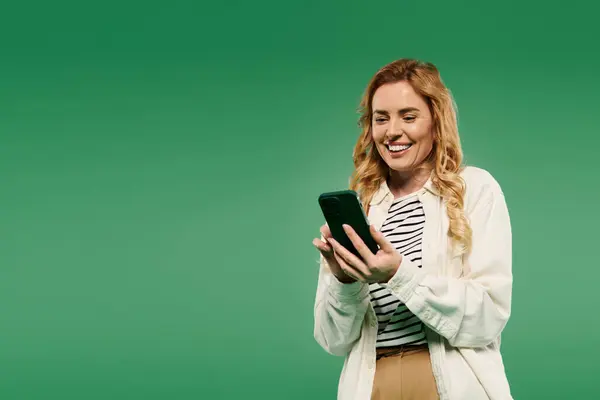 A cheerful woman dressed casually smiles while engaged with her smartphone against a vibrant green background. — Stock Photo