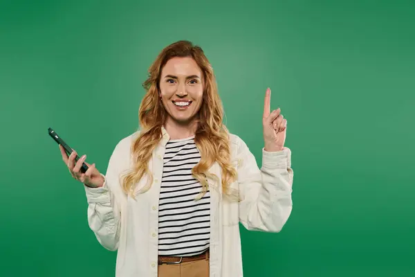 A cheerful woman with curly hair excitedly raises her finger while holding her phone, set against green. — Stock Photo