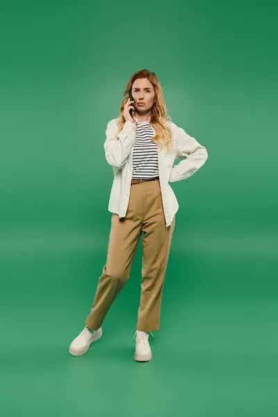 A confident woman with curly hair chats on her phone, dressed casually in a stylish outfit against a green backdrop. — Stock Photo
