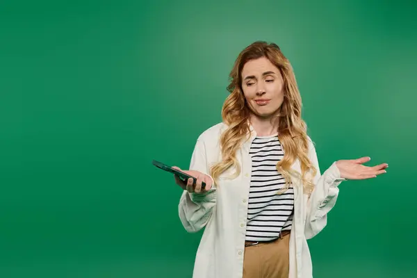 A stylish woman with curly hair engages playfully with her phone against a lively green backdrop. — Stock Photo