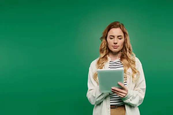 A woman with beautiful curly hair stands casually, focused on her tablet while surrounded by a vivid green background. — Stock Photo