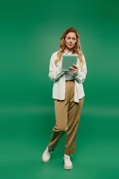 A beautiful woman with curly hair stands casually, engaged with a tablet, surrounded by bright green hues. — Stock Photo