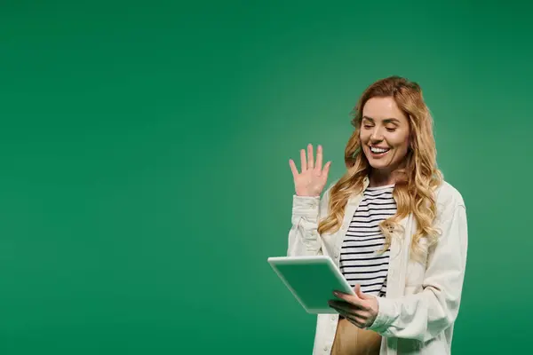A young woman with curly hair in casual attire joyfully waves while using a tablet on a vibrant green backdrop. — Stock Photo