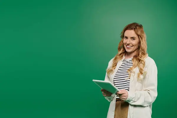 A joyful woman stands confidently, interacting with a tablet while dressed casually in a bright green setting. — Stock Photo