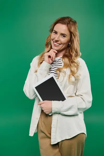 A smiling woman with curly hair stands confidently, holding a tablet and dressed in casual attire, radiating joy. — Stock Photo