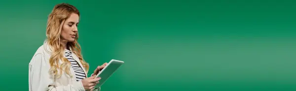 A beautiful woman in casual attire explores her tablet, captivated by the vibrant green backdrop. — Stock Photo
