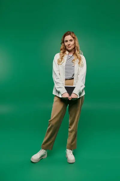 Una mujer alegre con atuendo casual se levanta contra un fondo verde brillante, irradiando confianza y estilo. - foto de stock