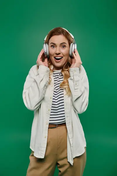 Une femme vivante aux cheveux bouclés exprime sa joie en portant un casque sur un fond vert vif. — Photo de stock