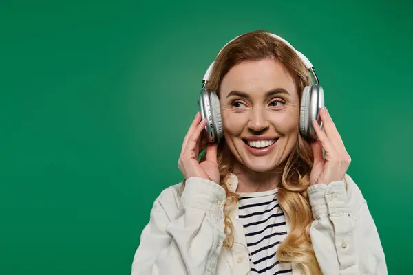 A cheerful woman with curly hair smiles broadly, wearing headphones and casual clothing against a green background. — Stock Photo