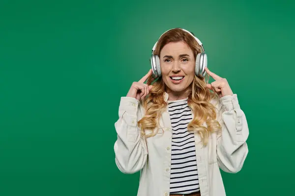 A happy woman with curly hair listens to music with headphones, smiling against a green background. — Stock Photo